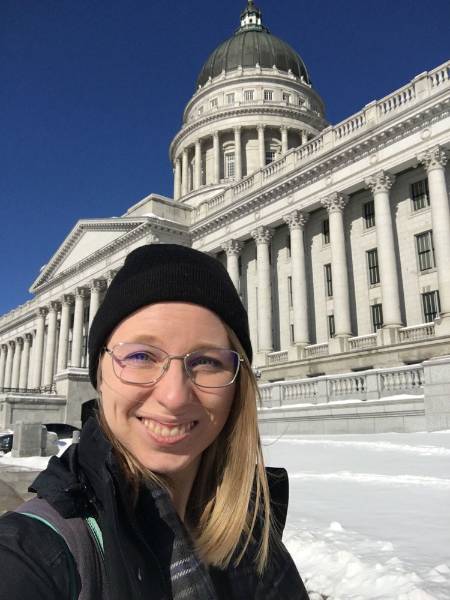 Anne stands in front of the Utah State Capitol Building on a sunny winter day. There's snow in the background and she's wearing a black hat and black jacket.