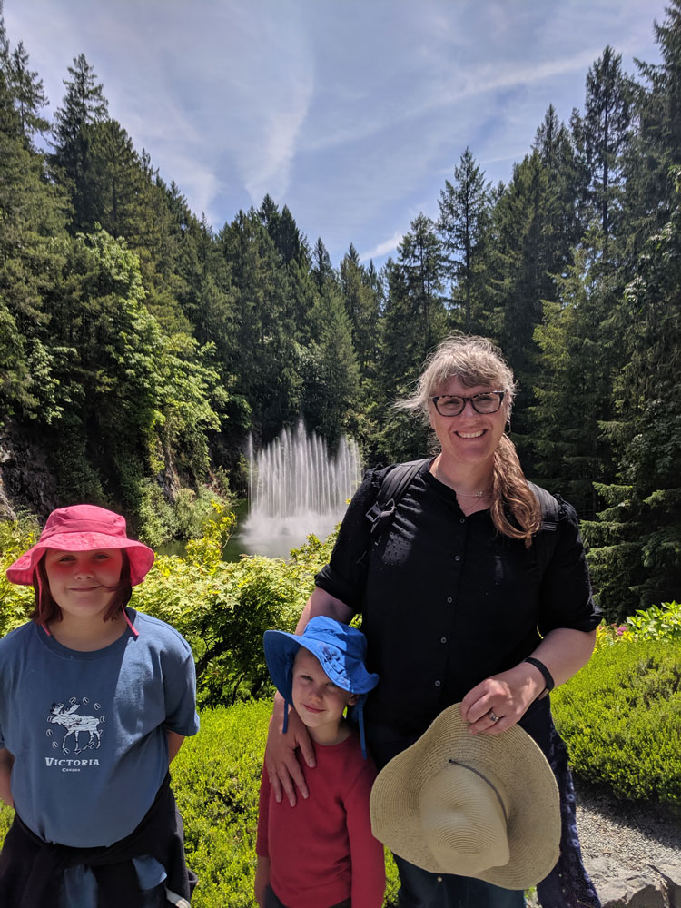 Danielle Endres stand with her two kids on a sunny day outside in front of a fountain and pine trees.
