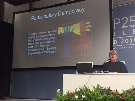 Danielle Endres gives a presenation at COP25, she sits behind a white podium with a projector screen behind her that says Participatory Democracy.