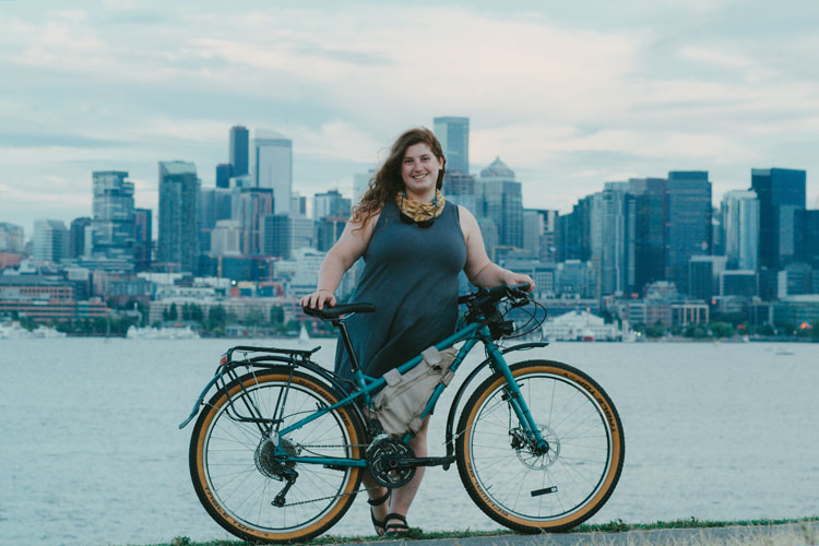 Kailey stands holding a gravel bike with a water and city scape behind her.