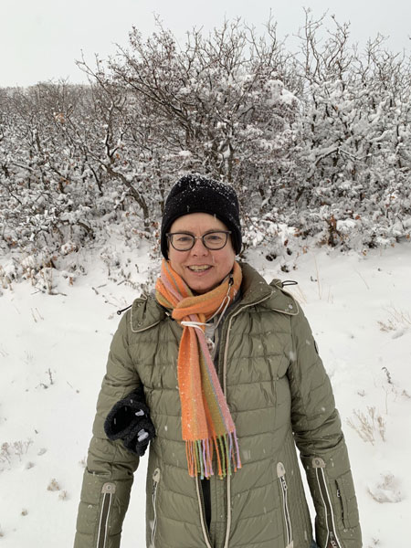Katharina stands in front of a snow covered hill wearing an olive green puffy and a multicolered scarf.