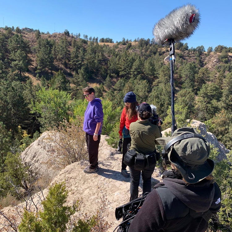 LaUra and Aimee being filed for a documentary. Four people stand on rocks surrounded by pine trees. Two hold camera gear, the other two stand looking in the distance.