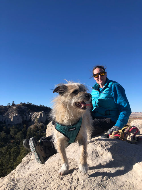 A white woman wearing a bright blue puffy jacket and sunglasses sits on a cream colored rock next to her small dog.
