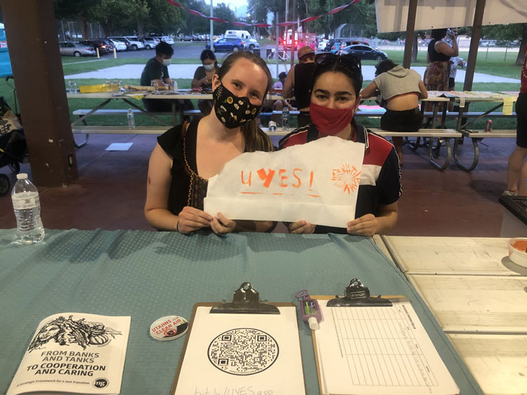 Maria holds a sign that says UYES with a student organizer. They sit at a picnic table under a pavillion.