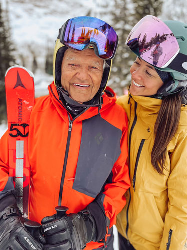 Ayja, a white woman with long brown hair, wears a mustard yellow ski jacket and mint green helmet with ski goggles. She has her arm around her grandfather, an older white man in an organge ski jacket holding red skis and wearing a helmet with goggles up. They are outside in the snow.