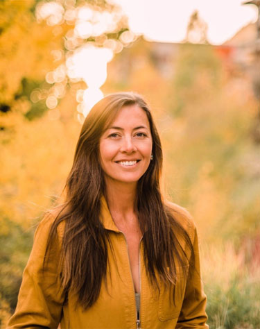 Ayja, a white woman with long brown hair, stands in front of blurred trees in a mustard long sleeve jumpsuit.