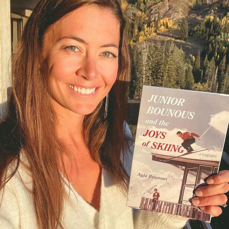 Ayja, a white woman with long brown hair wearing a cream sweater, holds a copy of her new book Junior Bounous and the Joy of Skiing.