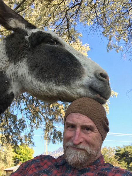 Eric stands with a donkey resting on his head. He wears a red flannel shirt and a brown beanie. The donkey is white and grey.