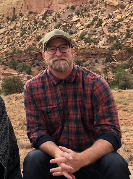 Eric sits in front of a sandstone landscape. He is wearing a red and blue flannel, an olive colored baseball cap, and glasses.