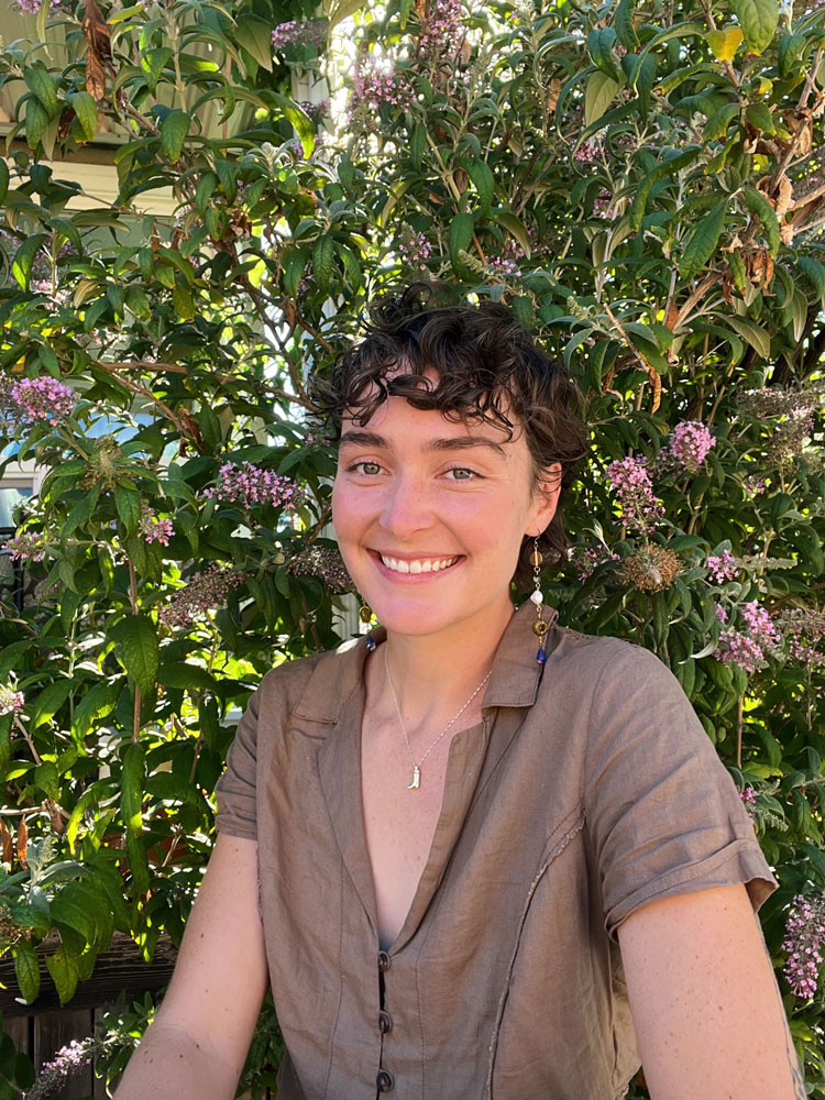 Madeline, a tall white woman with short brown hair, sits in front of bush with purple flowers. She's wearing a brown, collard v-neck blouse, dangling earings, and a necklace that has a cowboy boot charm..