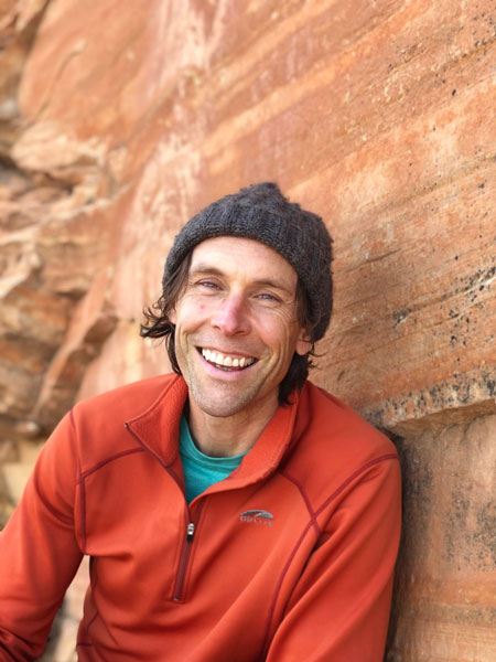Chris in an orange fleece and a beanie smiling in front of an orange sandstone wall.