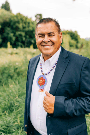 Headshot of Darren Parry, a Shoshone man, dressed in a white button up dress shirt, a blue suit jacket, and a traditional beaded Shoshone necklace. He is smiling and standing in front of a green field and forest blurred in the background.