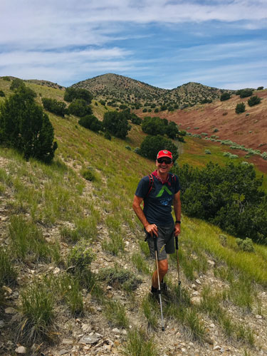 Jeff hiking in a green mountainside. He's wearing a blue tshirt, grey shorts, a red bandana around his neck, and a red U of U baseball cap. He is holding trecking poles.