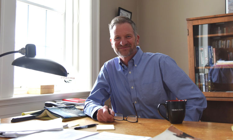 Jeff sitting at this desk, covered in papers and a mug. He's wearing a blue button up shirt.