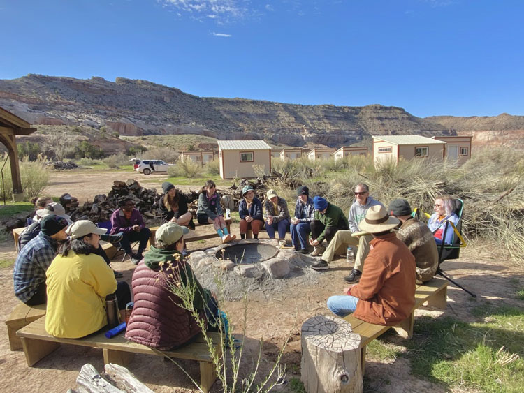 Students and Jeff sit in a circle on benches and camp chairs at the Rio Mesa Field Station in a remote red rock desert landscape. Sandstone cliffs are in the background.