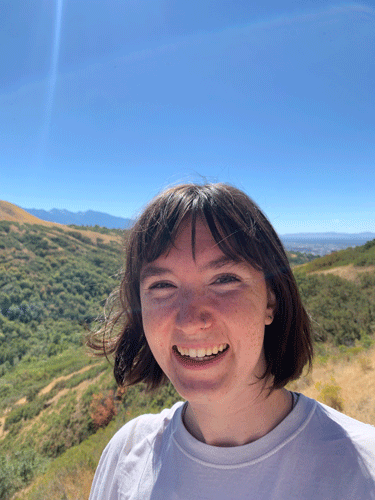 Headshot of Maggie, a white woman with freckles and short brown hair. She's wearing a white t-shirt. Green and brown mountain sides rolle in the background.