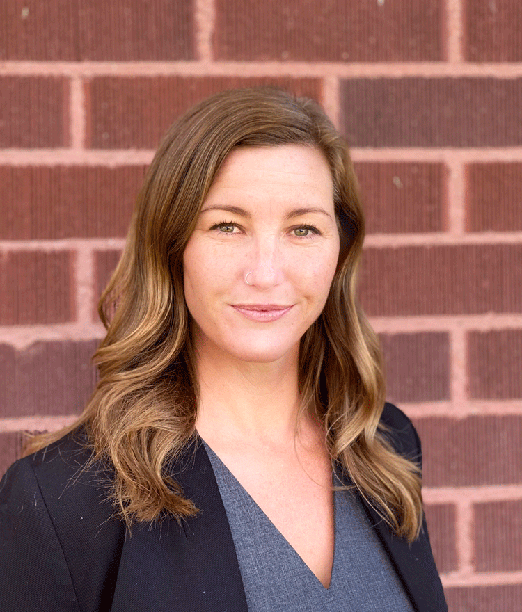 Headshot of Melissa Parks, a white woman with wavy long blonde hair wearing a grey blouse and a black blazer standing in front of a red brick wall.