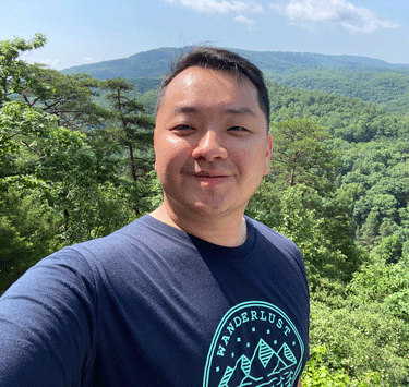 Headshot of Pheej in a blue t-shirt, smiling in front of a background of forested mountains.