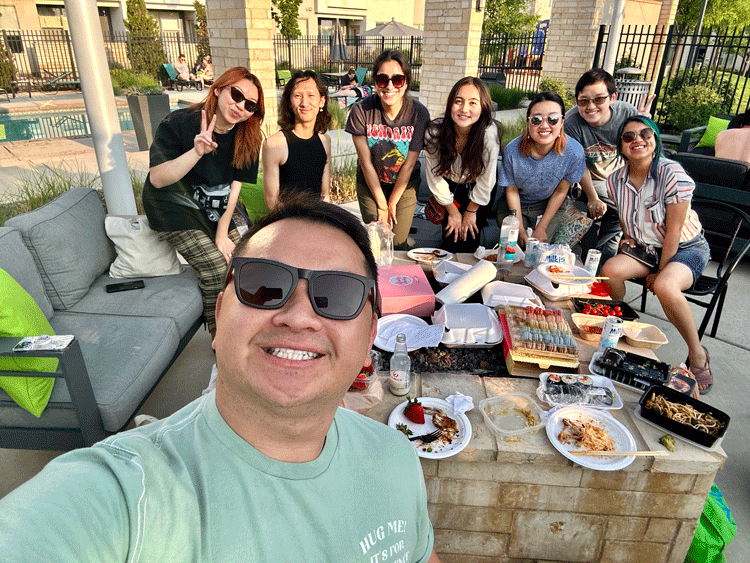 Pheej takes a selfie with a group of other Asian American young leaders. They sit outside near a pool around a table with food.