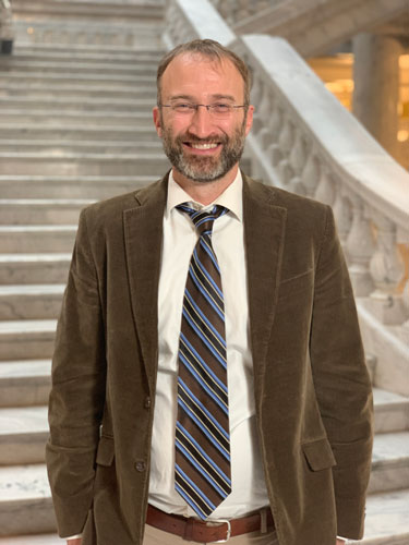 Ross, a white man with light brown hair and a light brown short beard, stands in front of the steps inside the Capitol Rotunda. He is smiling and wearing glasses, a white button up shirt, a brown cord suit jacket, and a tie with brown, blue, and white stripes.