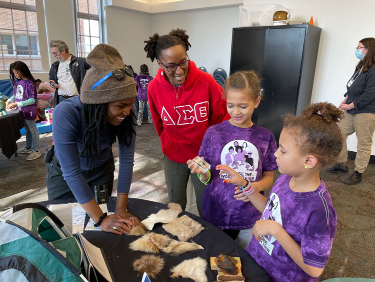 Sydney stands around a Black table that has animal furs and paw prints on it with two Black girls and a Black adult woman. They are all smiling as they look and touch the educational materials.
