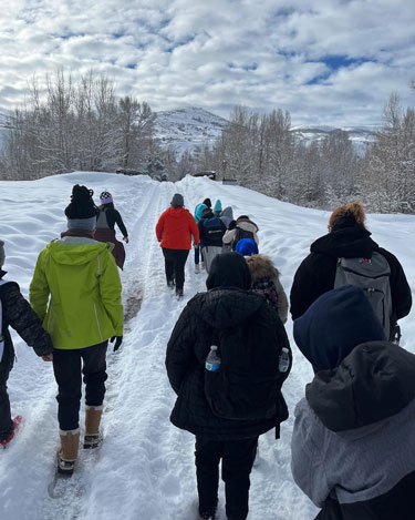 A group of young people walking on a snowy path, bundled in ski clothes. Image is of just the people's backs.