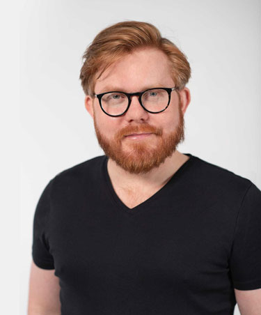 A headshot of Taylor, a white man with short red hair, a red beard, and glasses. He's wearing a black v-neck short sleeved shirt. Photo has a white background.