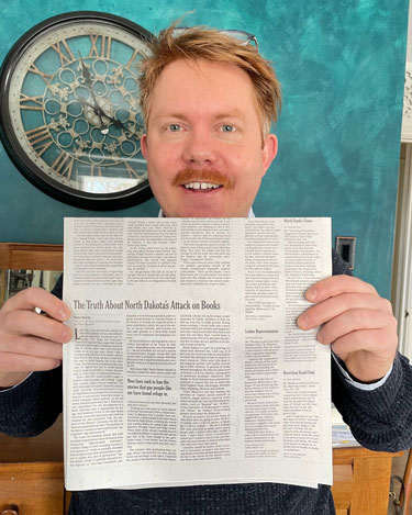 Taylor Brorby holds up a copy of The New York Times Opinion page with the article he wrote. Taylor is a white man with think, short red hair. Glasses are pushed on his head, he's wearing a grey sweater over a white collared shirt and standing in front of a blue wall with a big old fashioned looking clock.