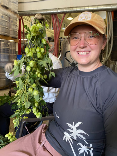 Tessa, a white woman with glasses and brown hair smiles while holding a branch of hops. She is wearing a beige hat, grey long sleeved shirt, and pink pants.