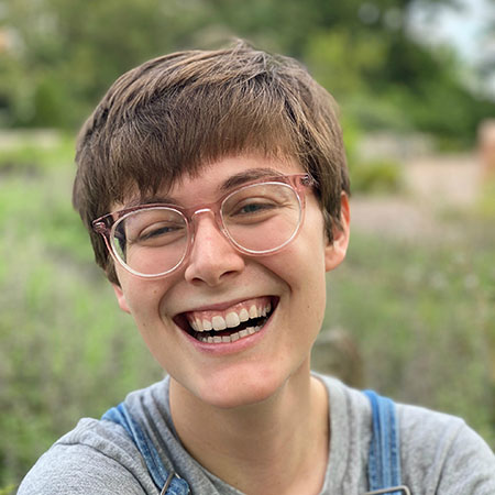 Natalie, a white woman with glasses and short hair, smiles in front of a blurred garden scene. The image shows shoulders up and she is wearing a grey shirt and denim overalls. 